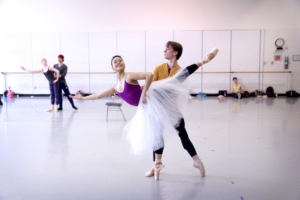 Angelica Generosa performs an arabessque elong\u00e9 on pointe while her partner stands behind her holding her waist and with his left leg in tendu. She holds her left hand on her hip and extends her right arm out to the side with her palm up. Angelica wears a purple leotard, black tights and a white Romantic tutu while Kyle wears a yellow shirt, black tights and tan slippers.