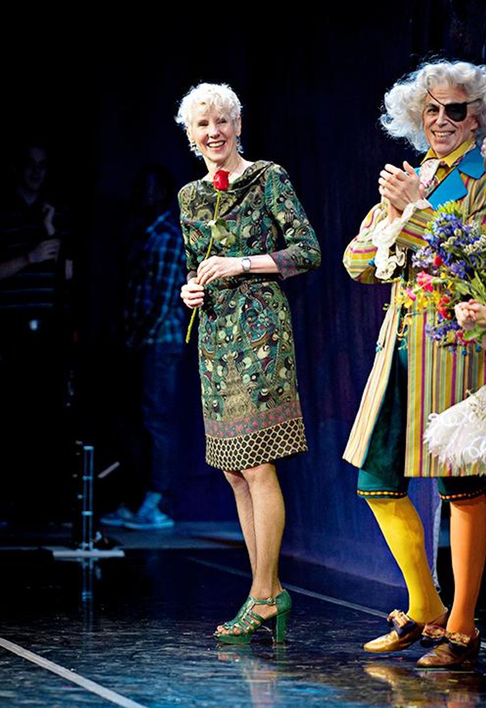 Victoria Morgan is shown from the side standing on stage right, turning to smile at a line of costumed dancers to her left during bows. She wears a patterned green dress with chunky green high heels and holds a red rose in her hand.