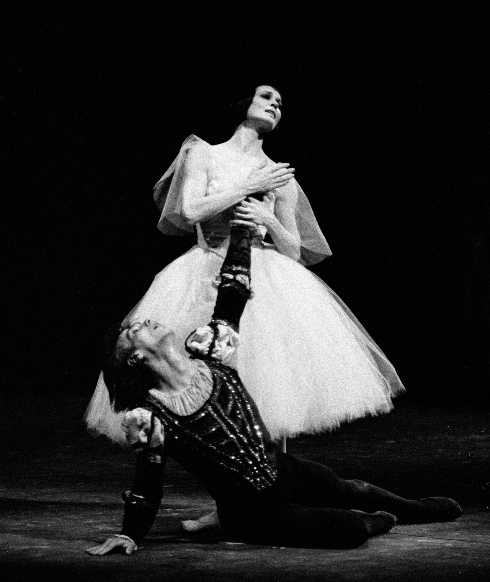 In this black and white photo, Carla Fracci stands above Gheorghe Iancu, who lies in a heap on the stage floor, during a performance of Giselle. She grabs his left hand and holds it to her heart while he looks up at her desperately. She wears a white Romantic tutu while he wears a dark mediaeval-style prince tunic and dark tights.