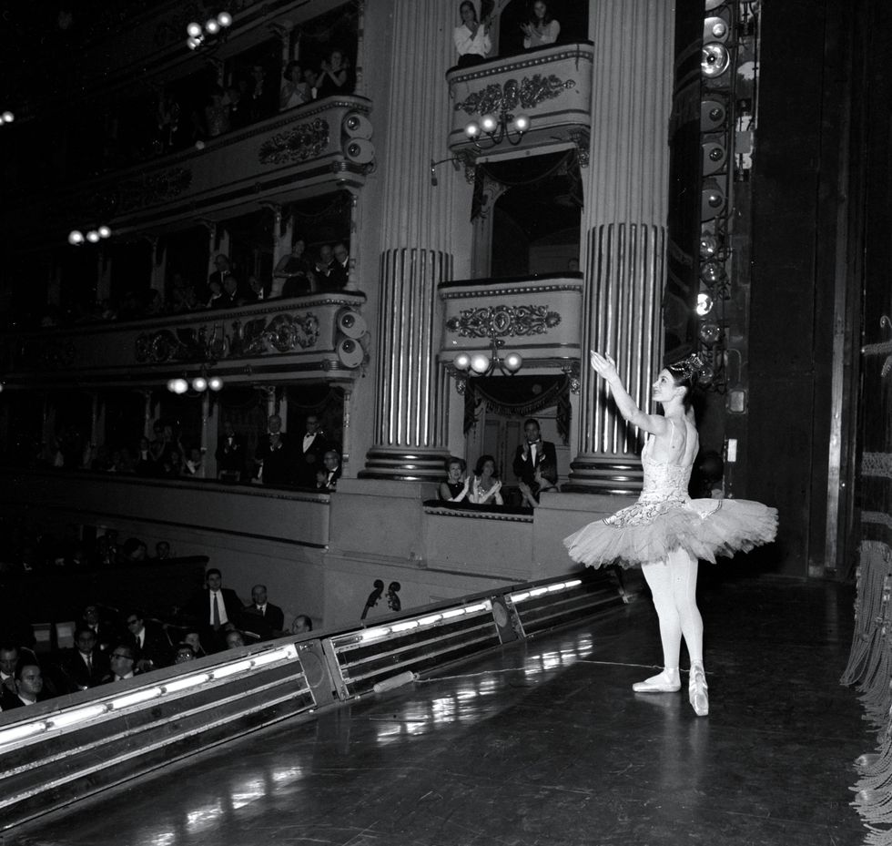 This black and white photo, taken from offstage left, shows Carla Fracci in a regal tutu, tights and pointe shoes taking a page bow onstage for an audience.