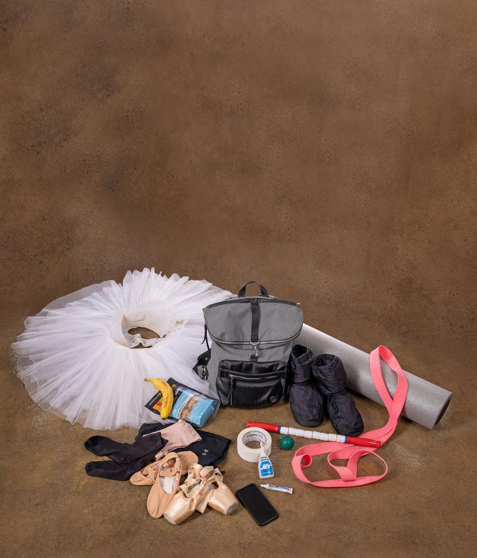 A tutu and dance bag rest on the floor, surrounded by pointe shoes, ballet slippers, booties, a foam roller, snacks and various first aid supplies.