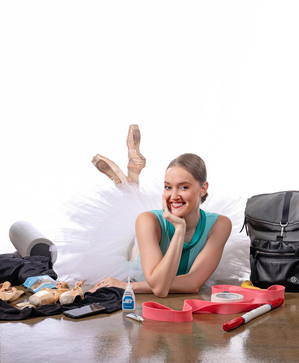 Sarah Lapointe lays on her stomach and props herself up on her right hand, tilting her head to the right and smiling towards the camera. She wears a green leotard, white tutu and pointe shoes, and is surrounded by various dance supplies.