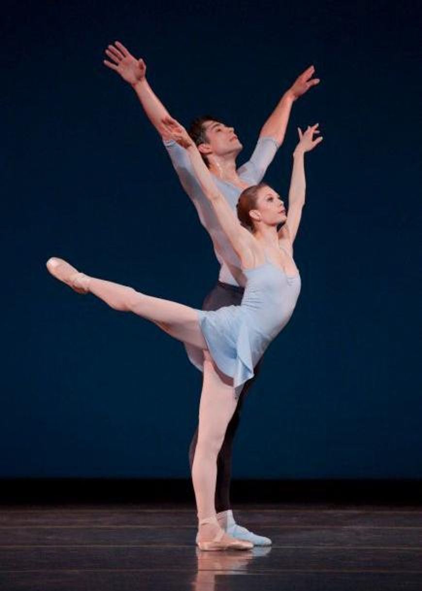 Gavin Larsen\u2014in a light blue leotard and skirt, pink tights and pointe shoes\u2014stands on her left leg with her right leg in a high arabesque and holds her arms up in a V-shape. Behind her, Artur Sultanov stands in sixth position with his arms in the same V-shape, and wears black tights, a blue T-shirt and blue socks and ballet slippers.