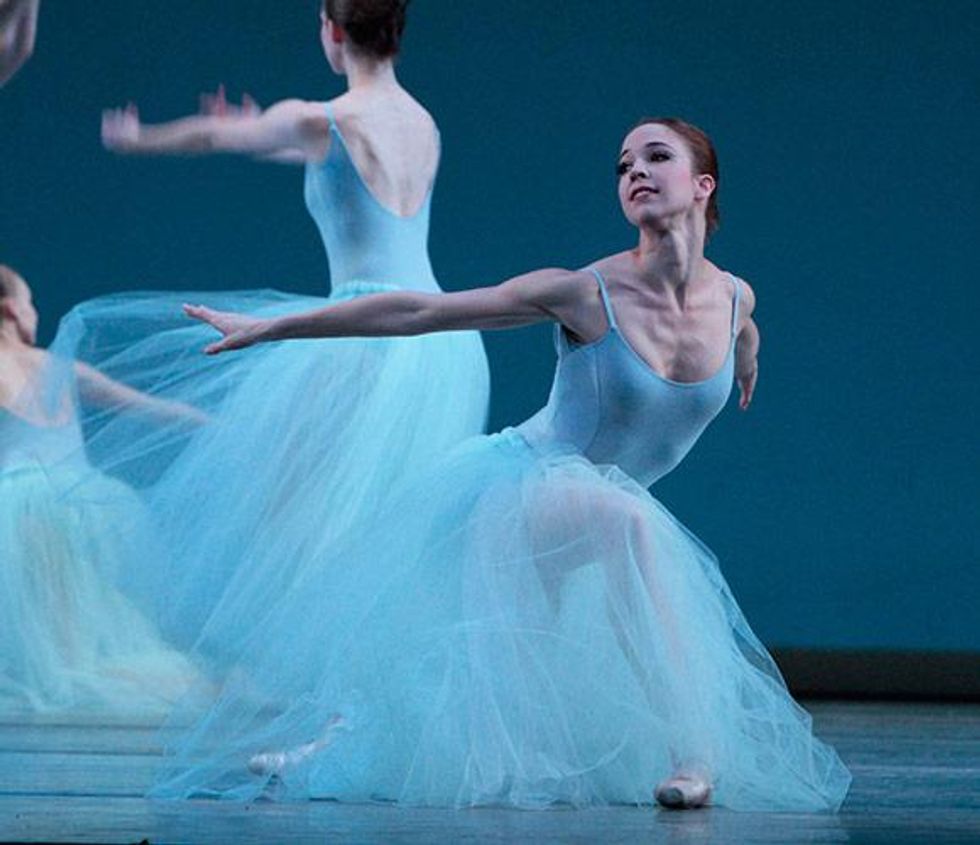 Lucy Van Cleef kneels on her left knee and reaches both arms back, looking over her right shoulder, during an onstage performance. She wears a light blue costume with a long tulle skirt, pink tights and pink pointe shoes.