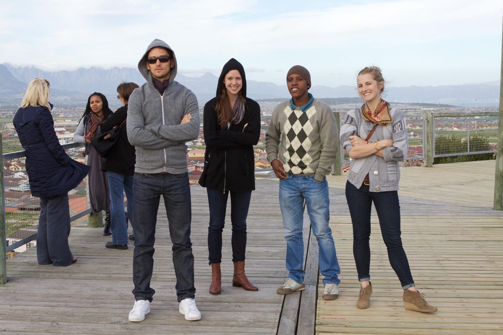 A group of two men and two women stand on a wooden deck overlooking a town and smile towards the camera. They wear jeans and assorted jackets, sweaters or sweatshirts.