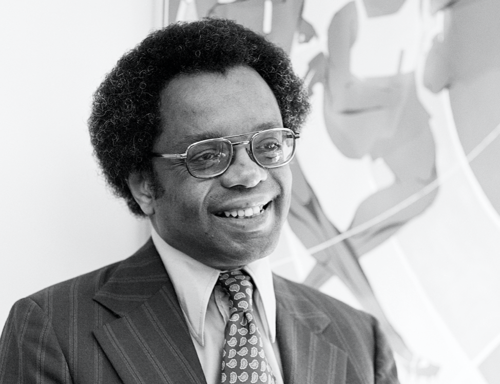 Black and white image of an academic looking black man in a striped suit with a small afro and aviator style glasses, the man is smiling and looking out of frame.