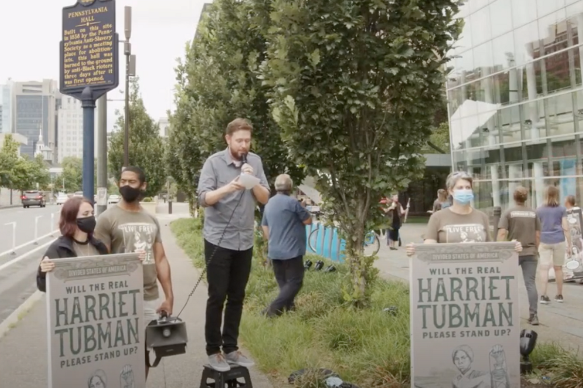 Bearded white guy stands on stool next to signs reading "Will The Real Harriet Tubman Please Stand Up"