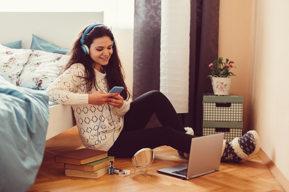 Girl sitting in her room on a smartphone listening to music with headphones and laptop opened on the floor