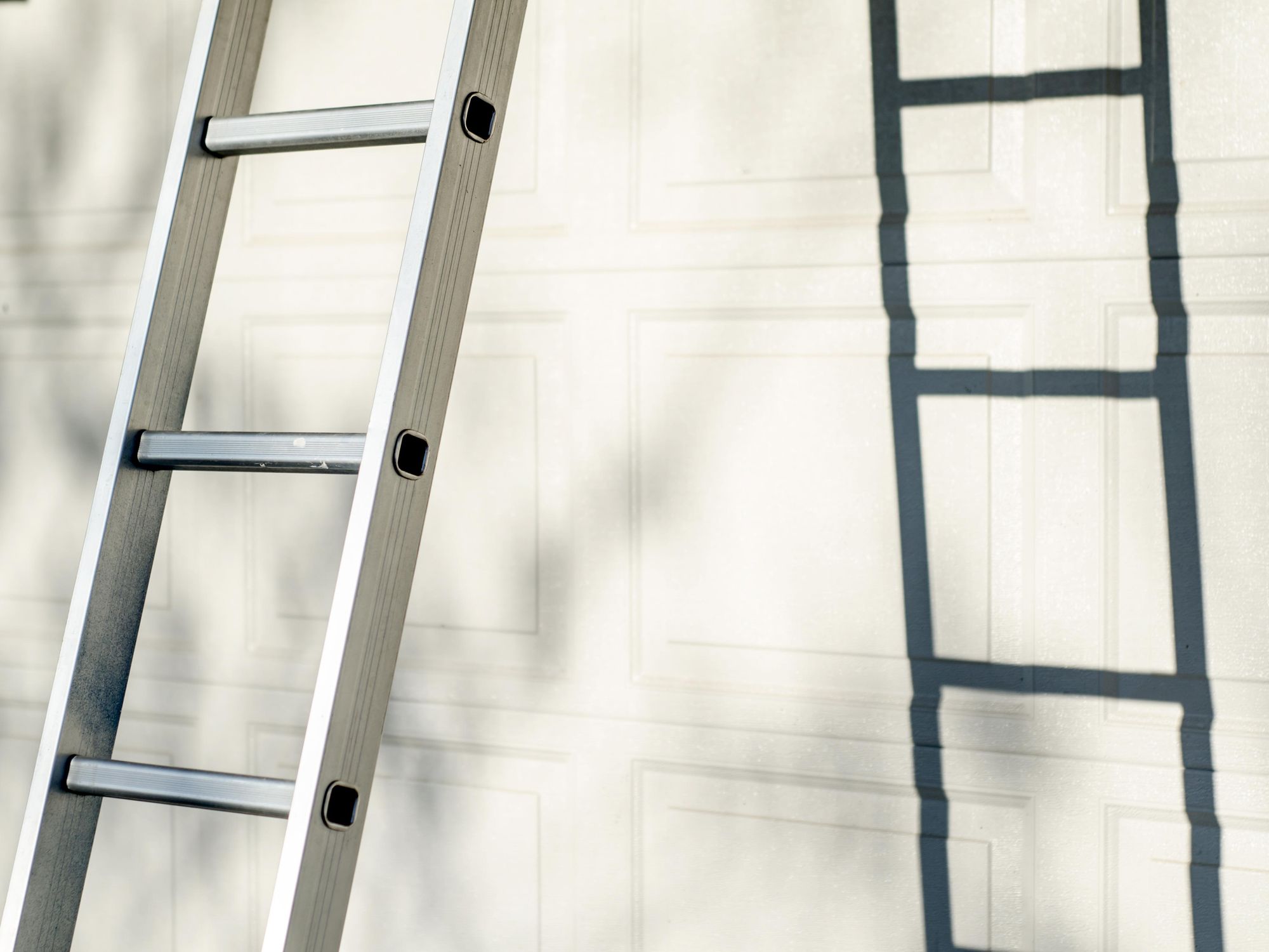 Close-up of an aluminum extension ladder leaning against a garage door, casting a shadow on the door panels