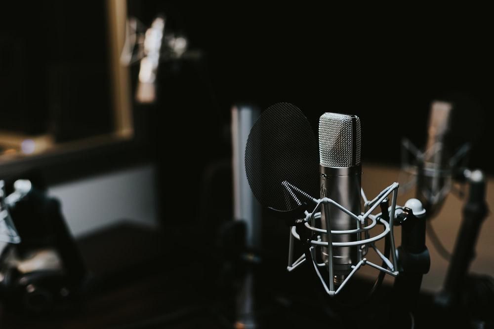 A dark blurred background with a silver microphone in the foreground. Looks to be a recording studio set up.
