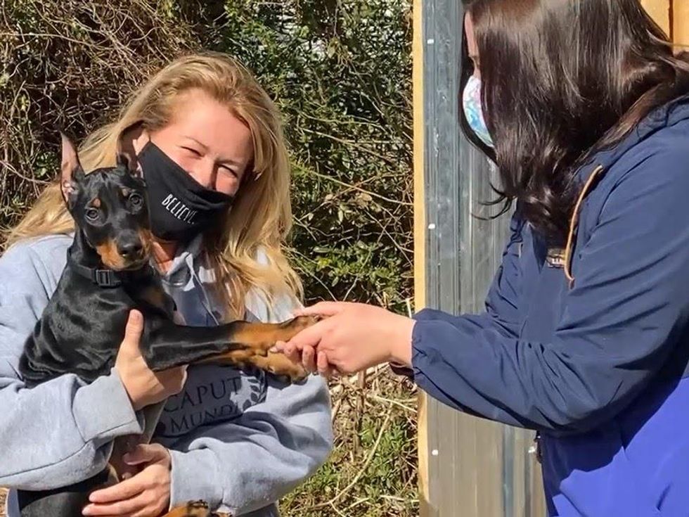 A woman holding a small black dog and looking at the camera is greeted by Jill Gonsalves