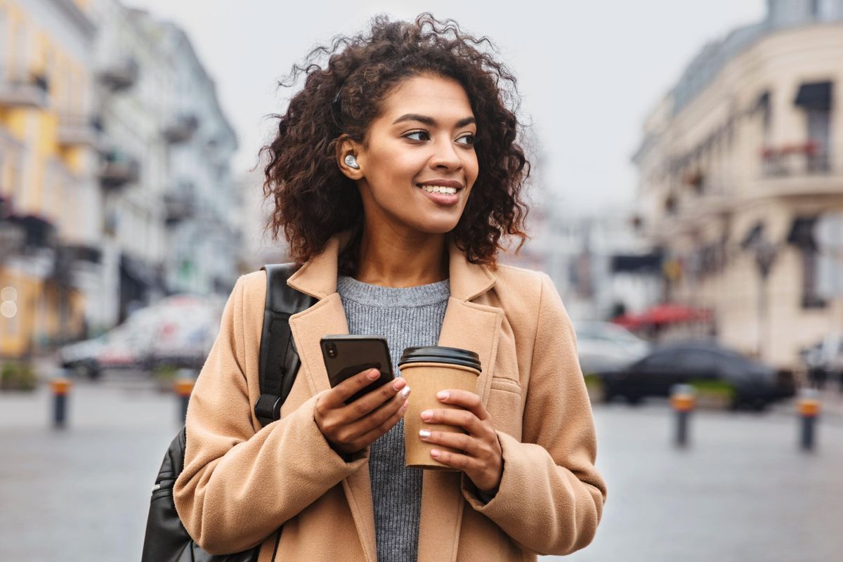 Woman wearing Technics earbuds and holding a coffee mug and smartphone