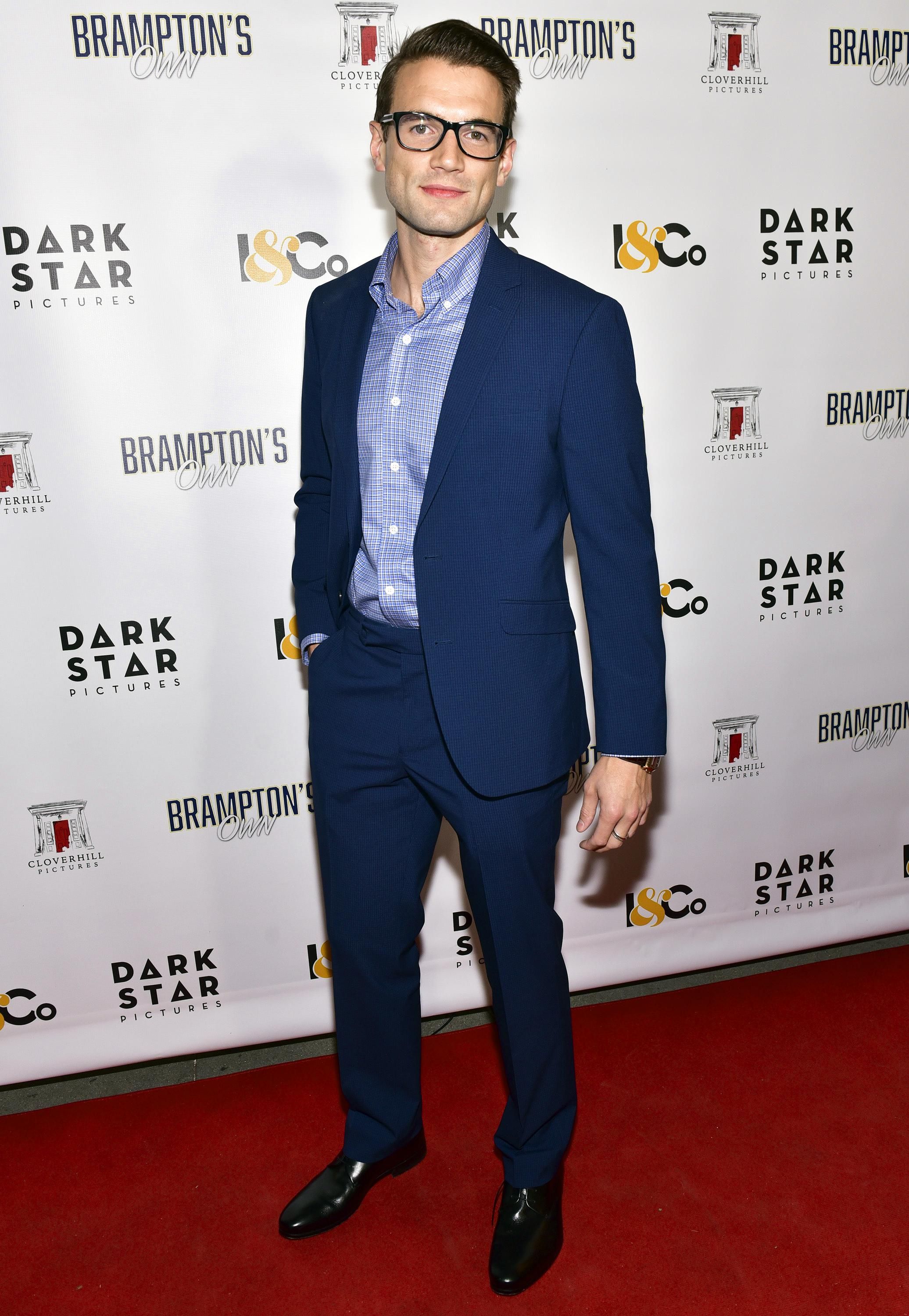 Alex Russell attends a red carpet event wearing navy blue suit and a checked blue shirt open at the collar