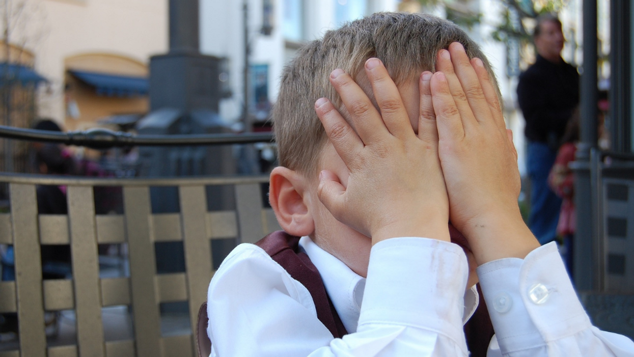 A young boy with light skin and short blond hair, wearing a long-sleeved white dress shirt and dark red suit vest, covers his face with his hands.