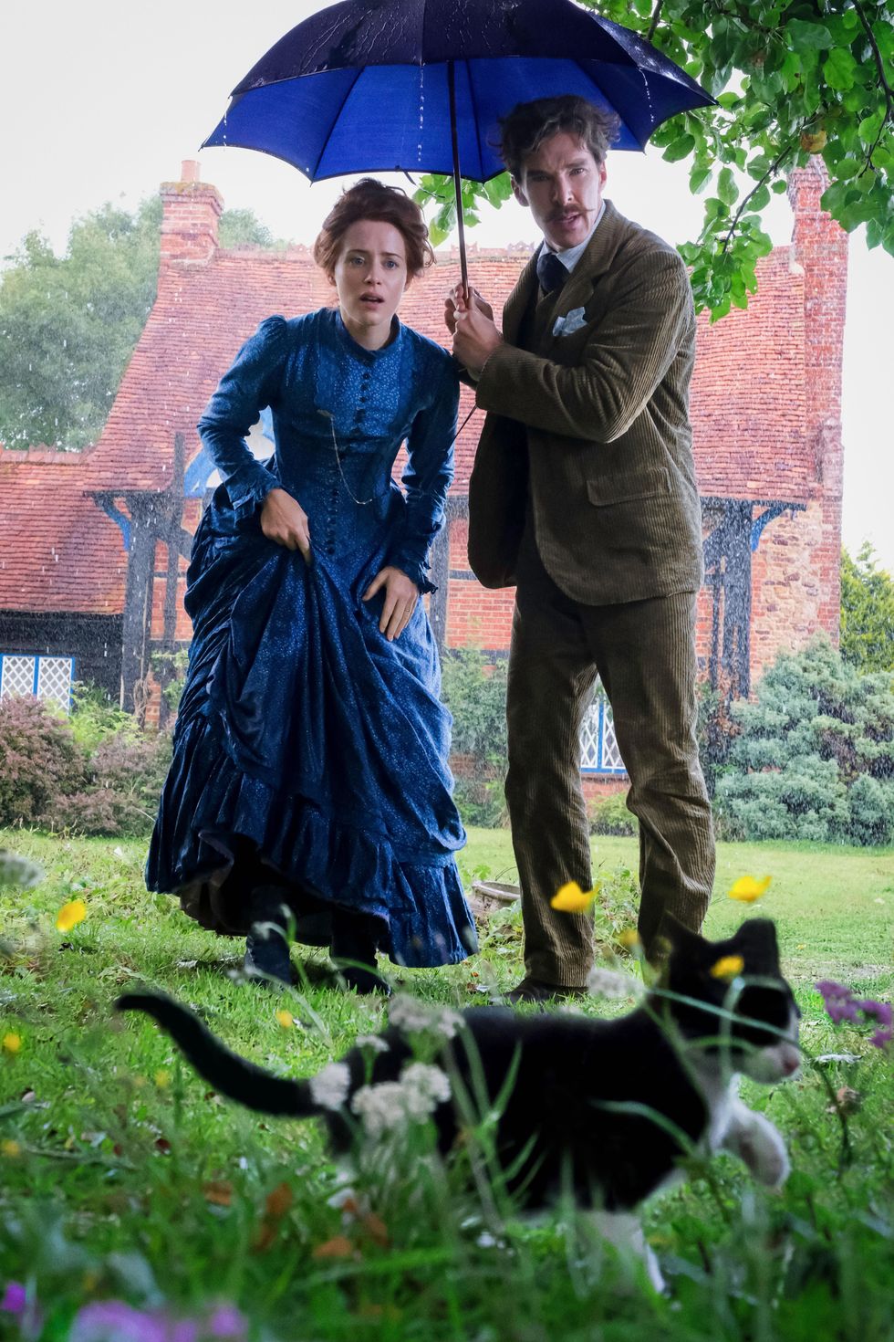Claire Foy (left) and Benedict Cumberbatch (right) discover a stray kitten on their lawn in the middle of the rain. Benedict holds a blue umbrella.