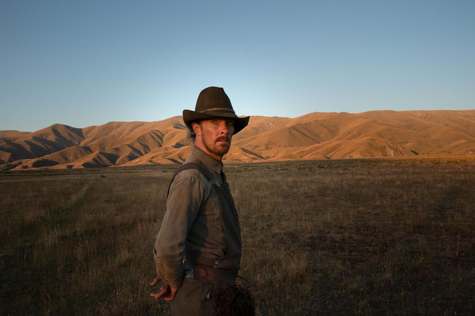 ​Benedict Cumberbatch as Phil Burbank in Netflix's "The Power of the Dog." He's wearing dark-colored work overalls a cowboy hat and a grey button-down work shirt. He looks off at the sunrise as light faintly bounces off his face. The mountains and plains are behind him.
