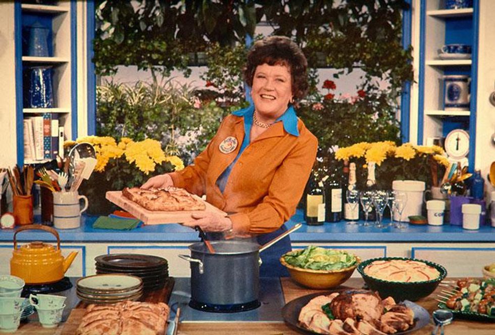 A photo of Julia Child on the set of her cooking show. She's surrounded by various dishes, but smiles and holds up one. 
