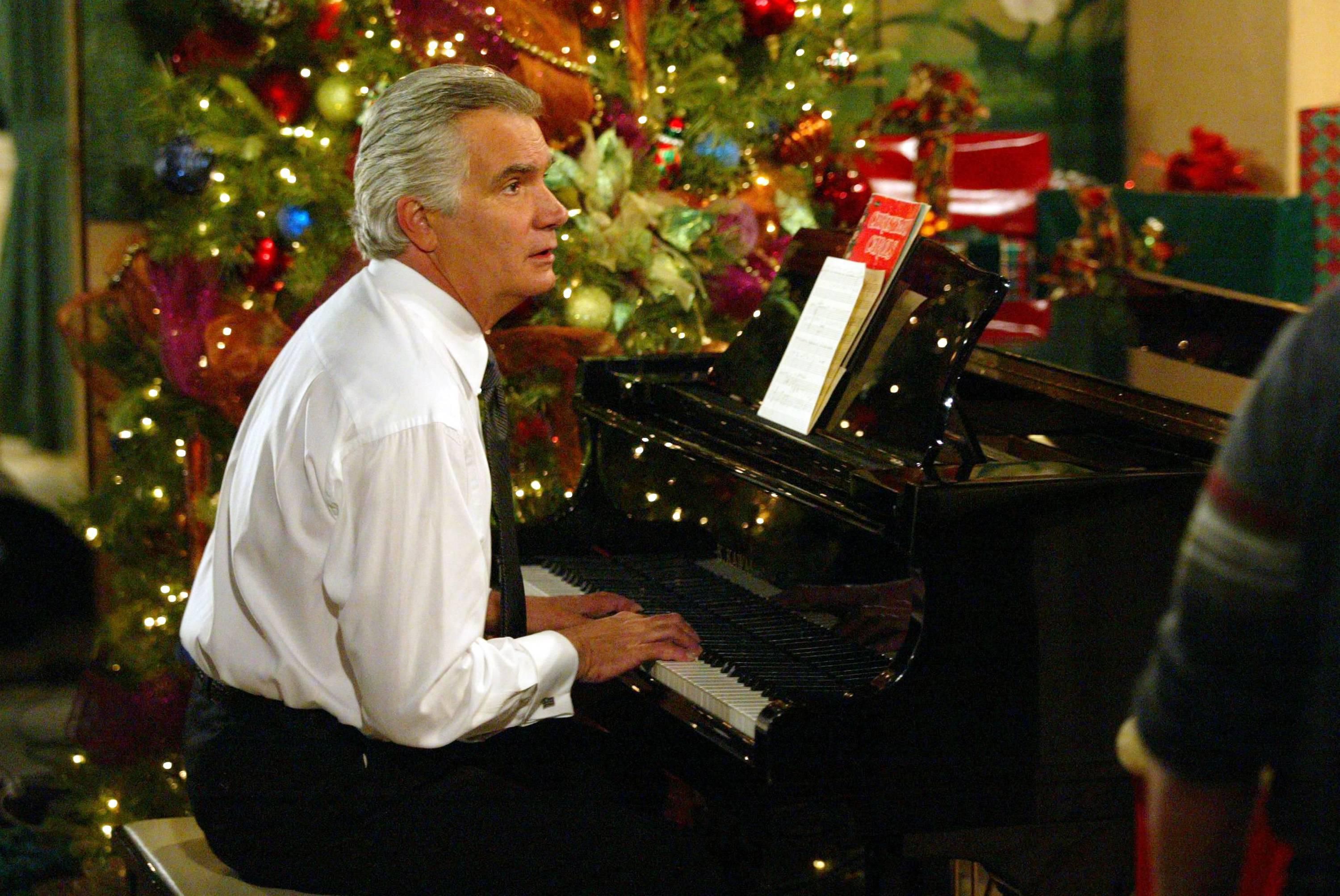 John McCook as Eric Forrester sitting down at a piano in front of the Forrester Christmas tree