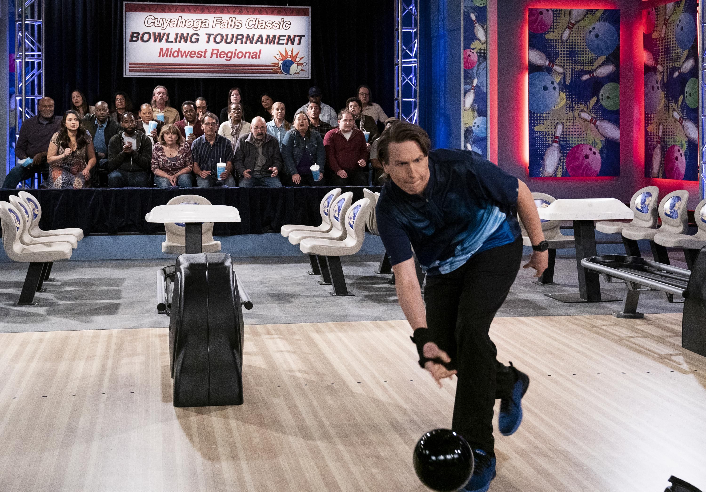 Pete Holmes as professional bowler Tom Smallwood throwing a black bowling ball with tournament onlookers in the background