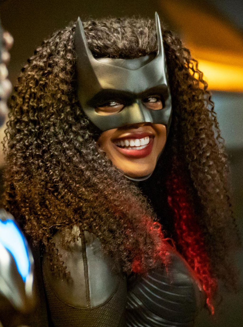 A smiling Javicia Leslie as Batwoman wearing her trademark bat mask and black action hero suit