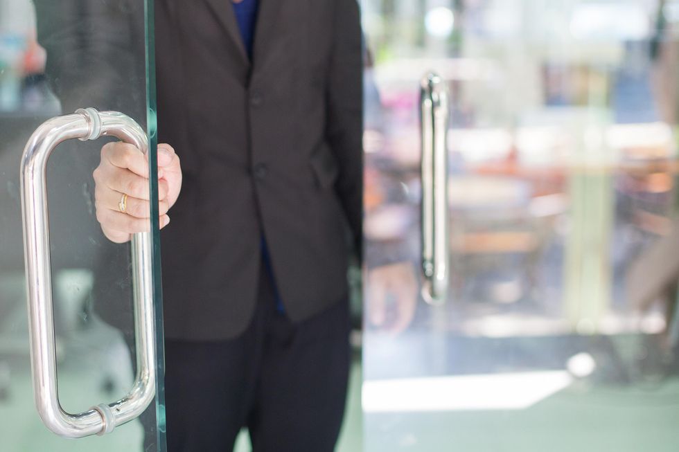 A man in a black suit holds a glass door open.