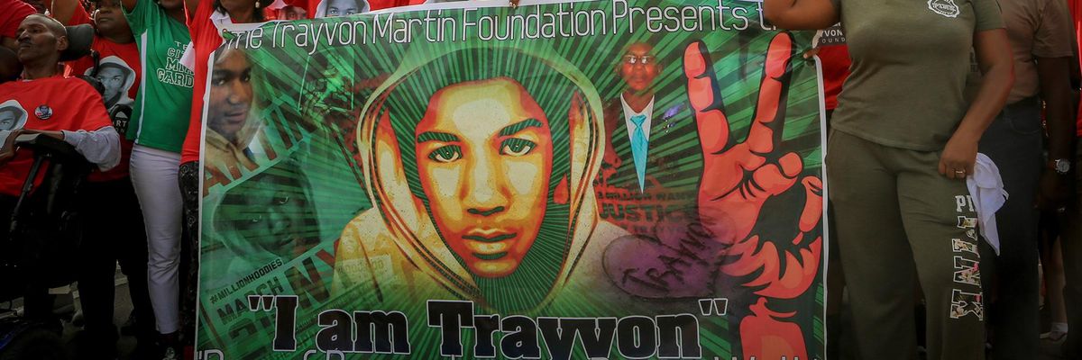 A group of people with right fists raised gather behind a banner showing images of Trayvon Martin and a hand drawn in a V-shaped peace sign