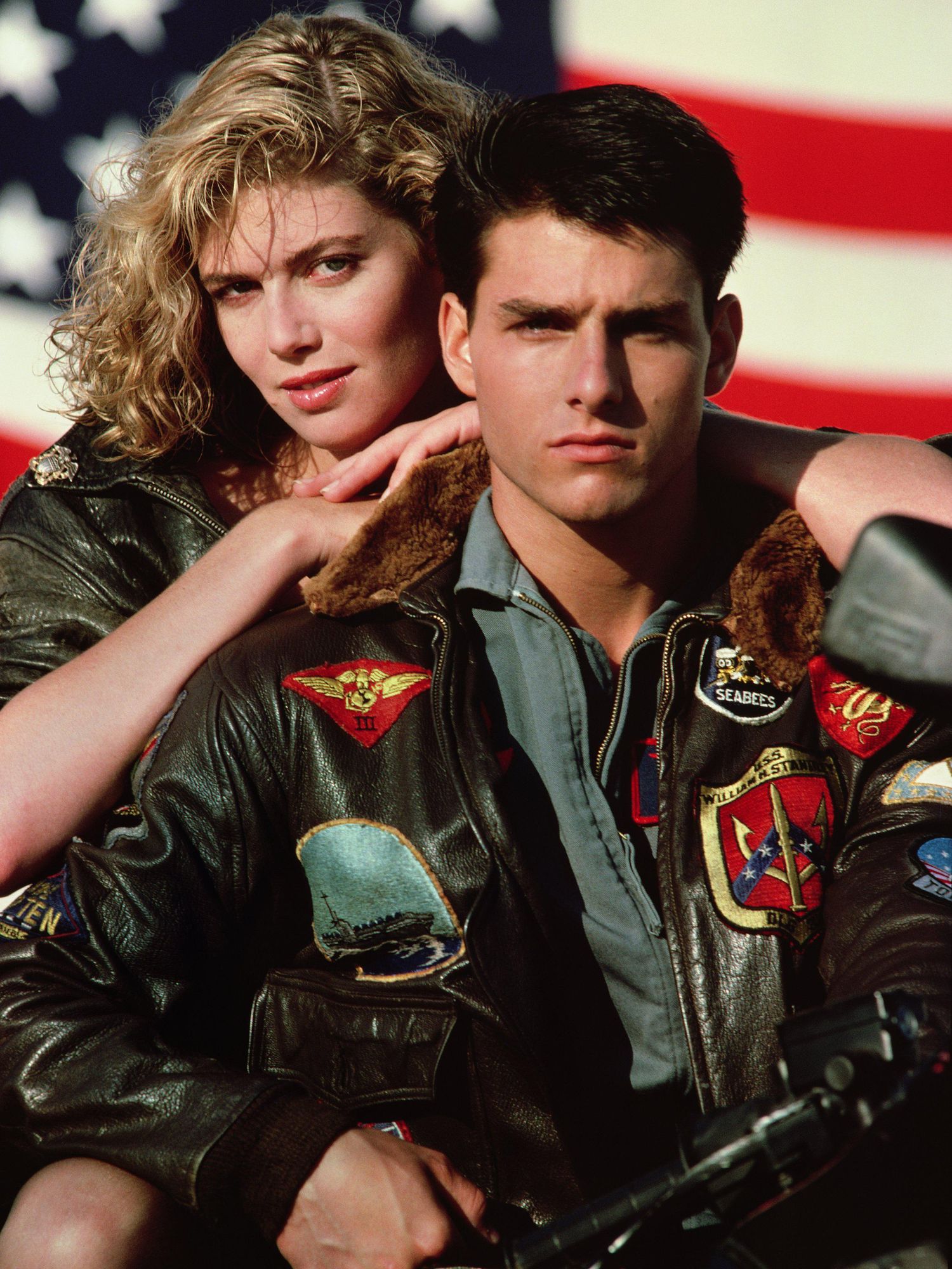 Kelly McGillis and Tom Cruise wear black leather bomber jackets with patches as they sit on a motorcycle in front of an American flag and she rests her hands on his shoulders