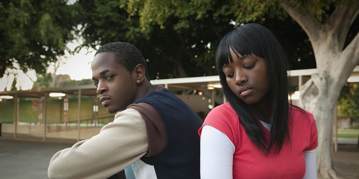 Black teen boy in a sweater vest and long sleeves sits and stares at the camera. A Black teen girl sits back to back with the boy, with hair down her shoulders and bangs. She looks down, sad