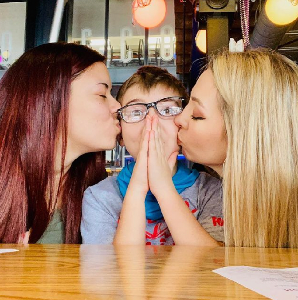 two women kissing a little boy wearing glasses on his cheeks