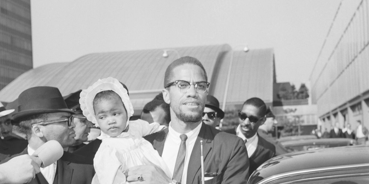 black and white image of Malcolm X in a suit, tie and glasses, stepping into a car while holding his toddler daughter who's wearing a bonnet, dress and dress shoes and shocks.