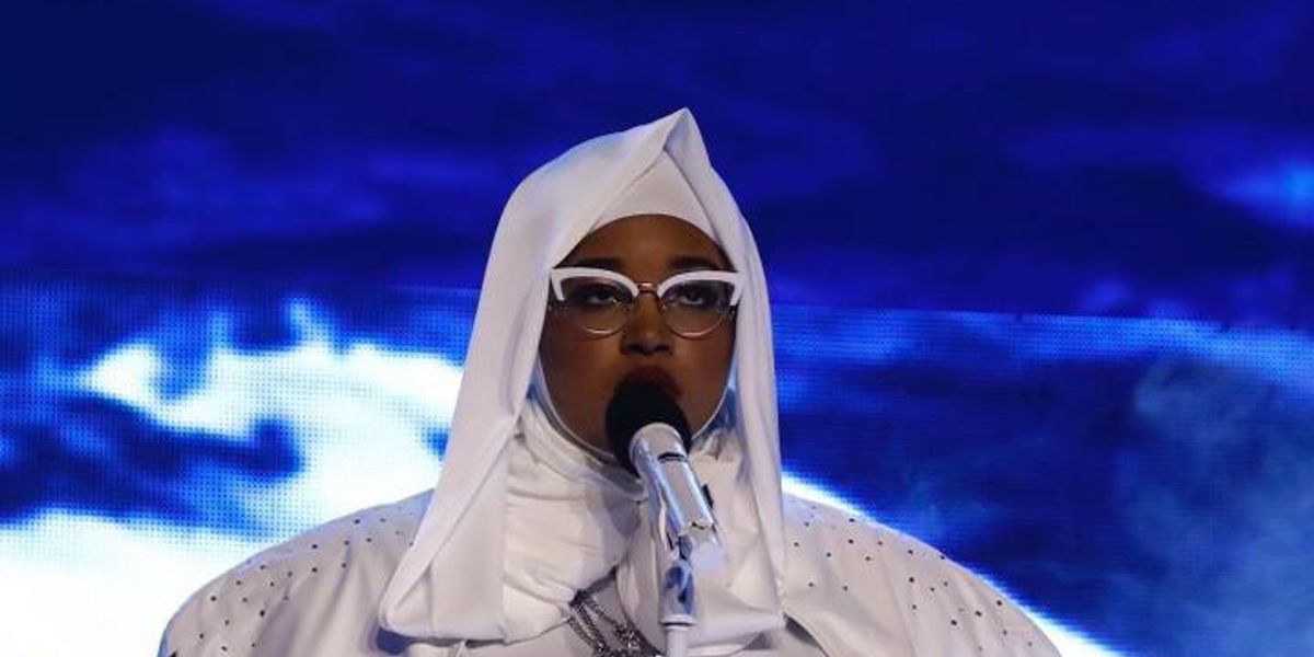 A Black woman in a white hijab, white cat-eyed glasses, white cape and outfit with rhinestones sings into a white microphone