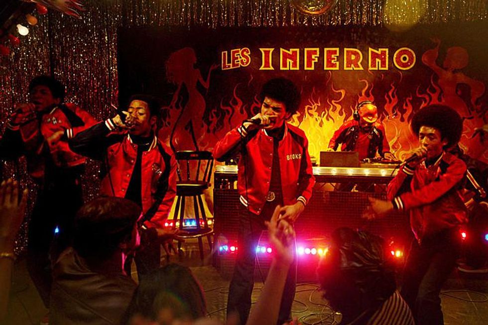 4 male hip-hop artists in matching red bomber jackets performing into mics on a fire-themed stage