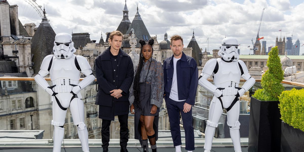 A Black woman, Moses Ingram, stands in a grey blouse and dark grey skirt with black sheer socks and black and gray boots in between actors Hayden Christensen, Ewan McGregor and two Star Wars Storm Troopers in London