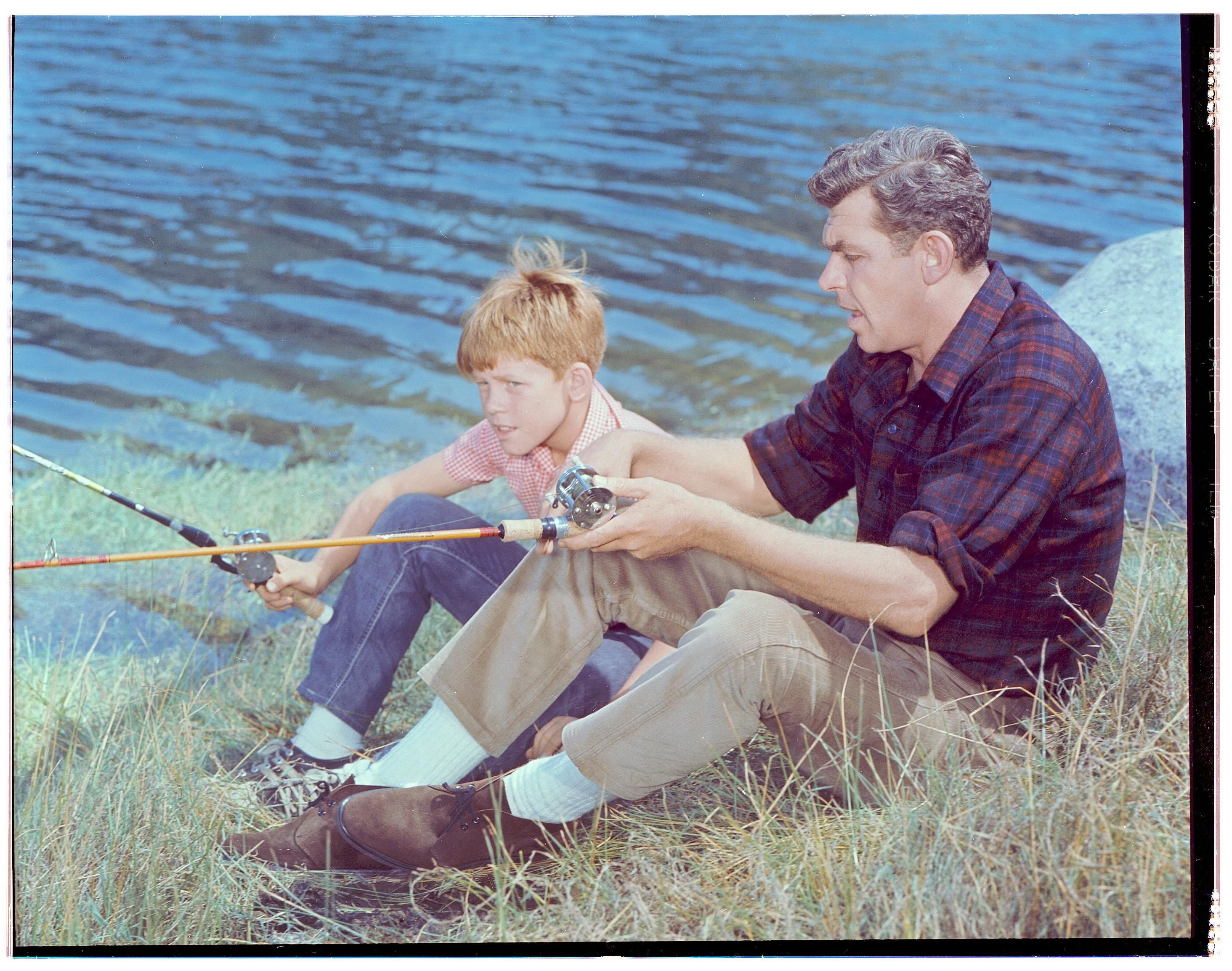 Andy Griffith and Ron Howard sit on the edge of a lake with their fishing gear in a scene from The Andy Griffith Show.