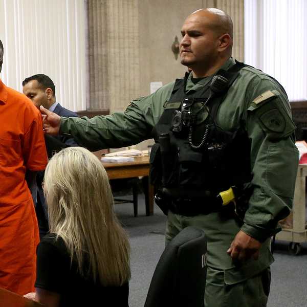 A Black man, R. Kelly, stands in a court room, wearing an orange jumpsuit with his hands handcuffed behind his back, accompanied by a police officer in a green uniform, bulletproof vest and gun.