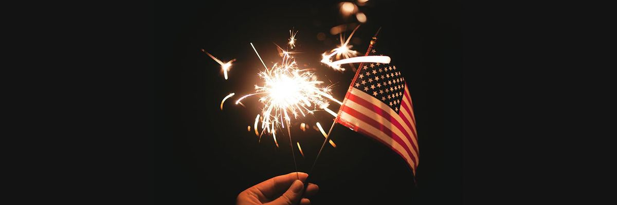 A hand holds up a lit sparkler firework and a small U.S. Flag against a dark background 