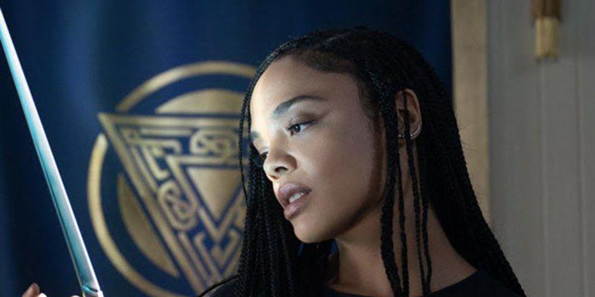 A Black woman, Tessa Thompson, in character as Valkyrie, wearing long black box braids, and looking admiringly at the glowing sword she's holding