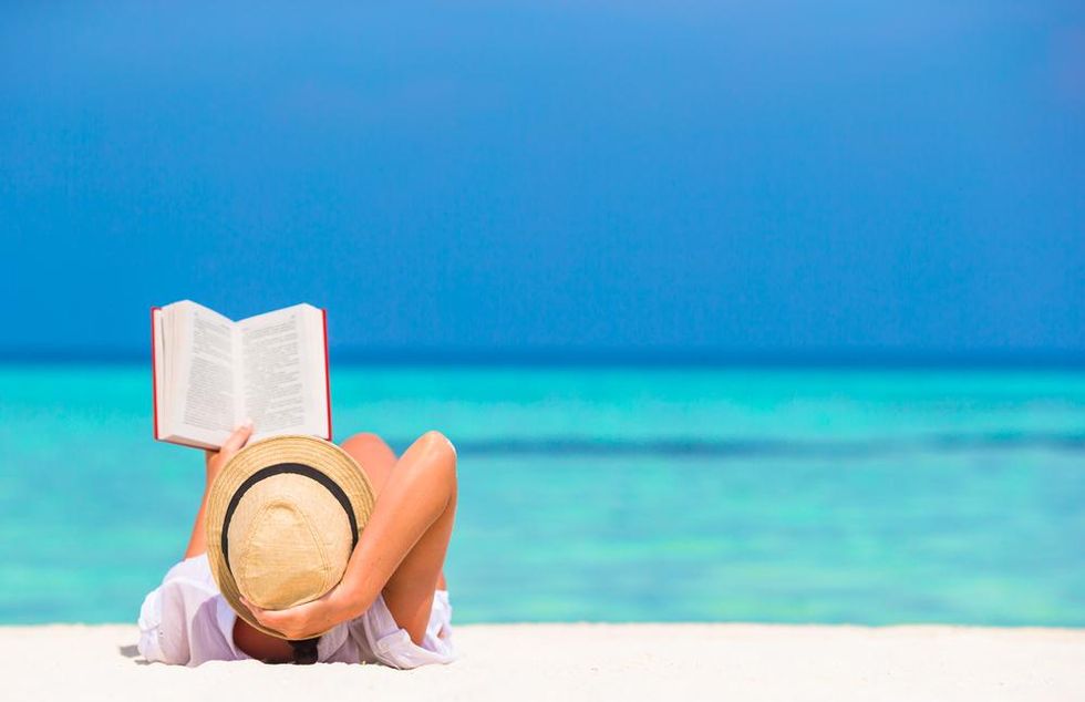 Woman in fedora lays on beach and reads a book