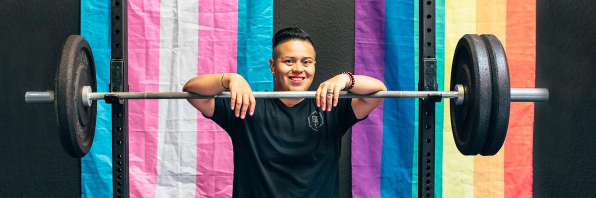 Fitness Coach Stands Facing Camera with hands over bar bell with weights on it in front of a Pride flag