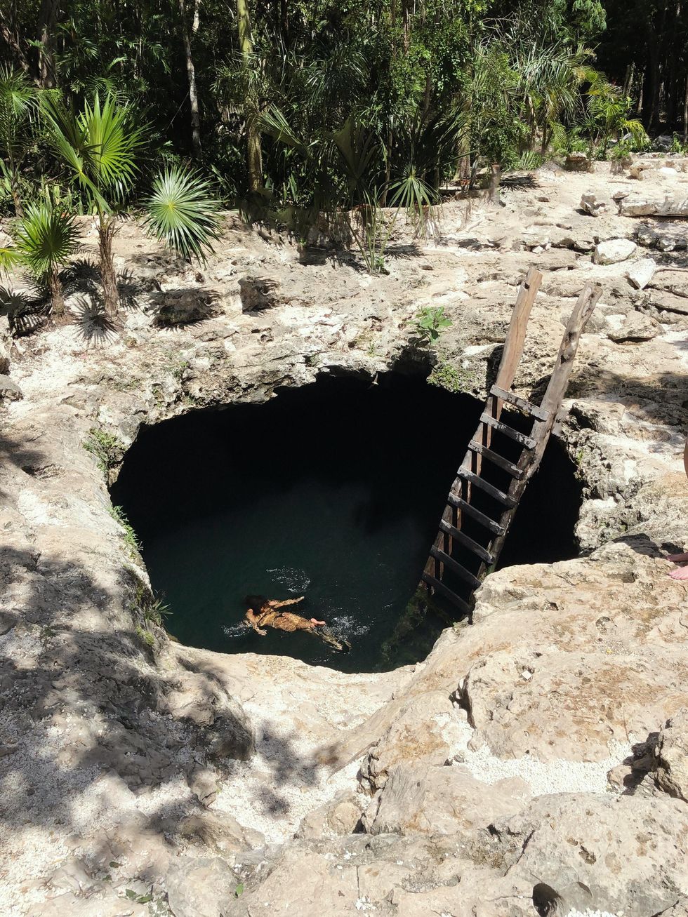 an aerial photo shows a woman swimming in a cenote with dark water surrounded by rocks and access by a ladder