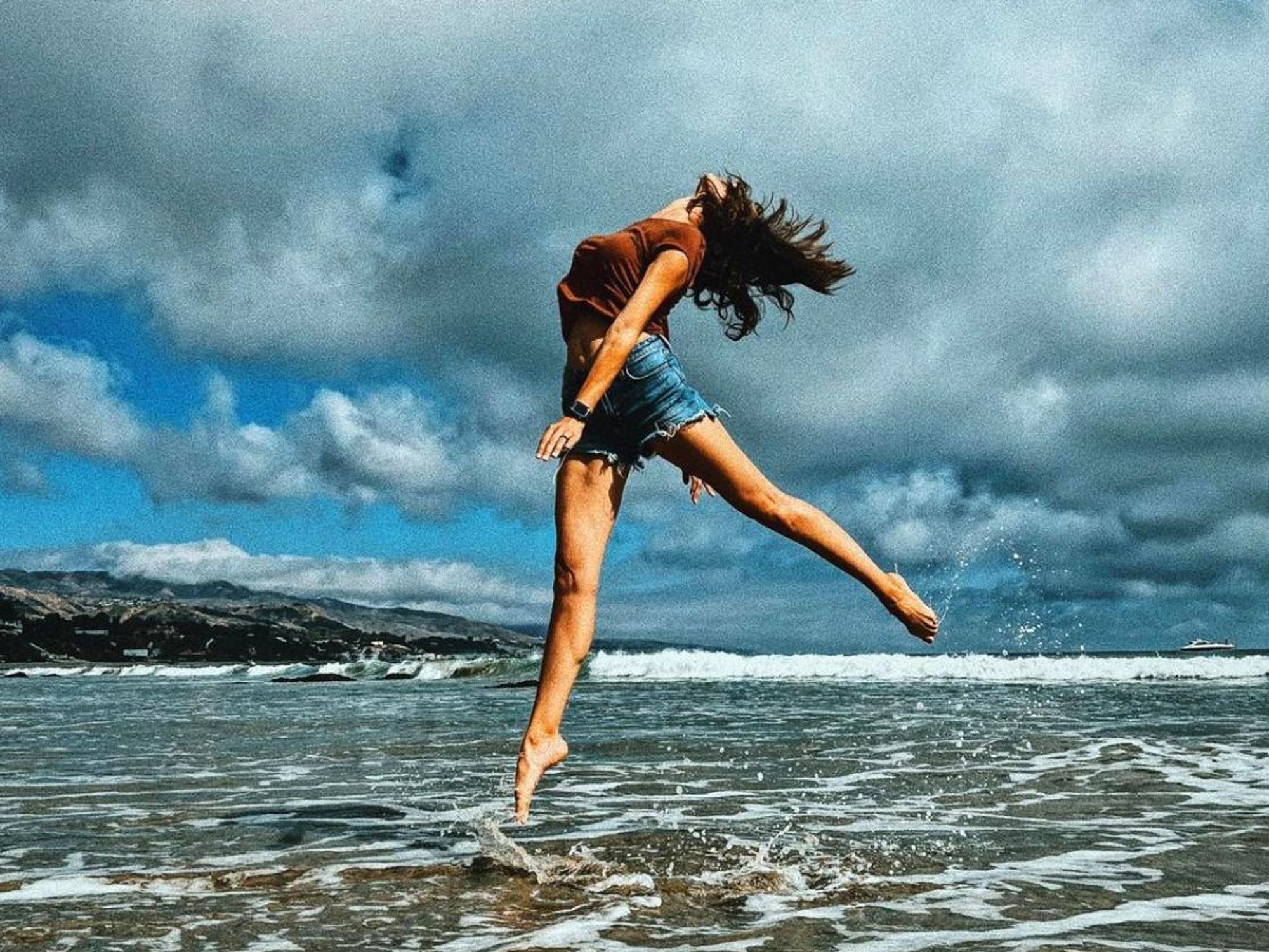 Daniela Ruah captured in a dramatic mid-leap as she kicks out from the surf on a beach with a clouded sky above.