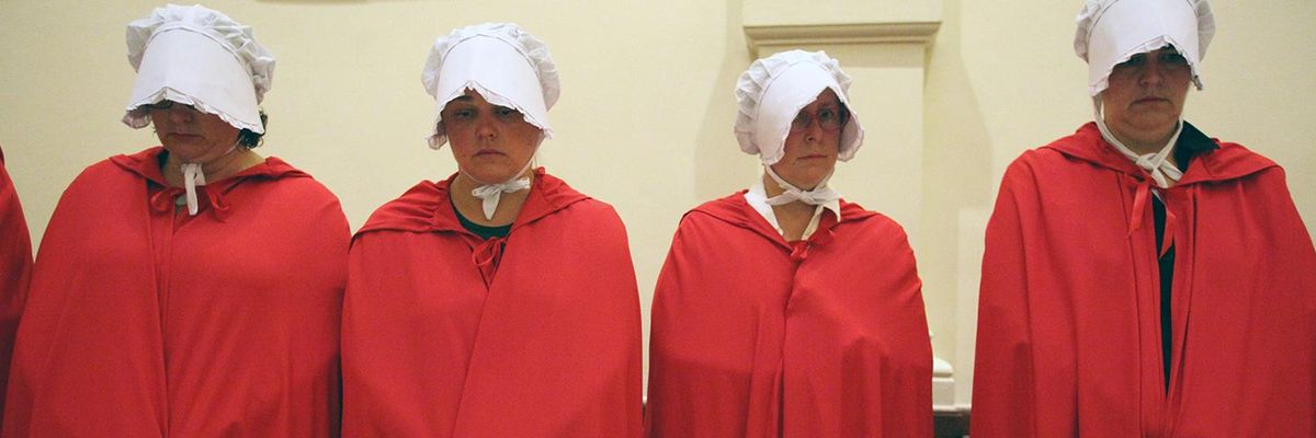 Four women dressed in red robes with white caps covering their faces look down solemnly