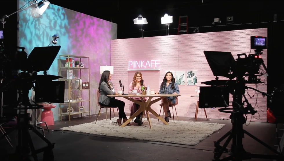 Three women sit at a table in a TV studio with microphones in front of them. The studio is decorated with a pink brick wall, bookshelf, rug, and a neon sign reading PINKAFE