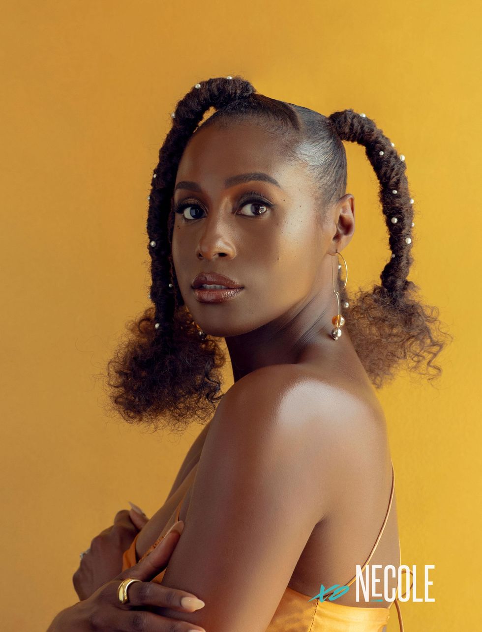 A Black woman, Issa Rae, stands to the side and looks over her shoulder, straight into the camera. Her mouth is partially open. Her arms are folded across her chest. Her hair is in four unicorn braids with curly ends and white pearls