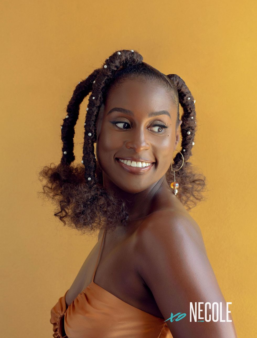 A Black woman, Issa Rae, stands to the side, looking off to the side over her shoulder and smiling. Her hair is in four unicorn ponytails with curly ends and each ponytail is studded with white pearls.