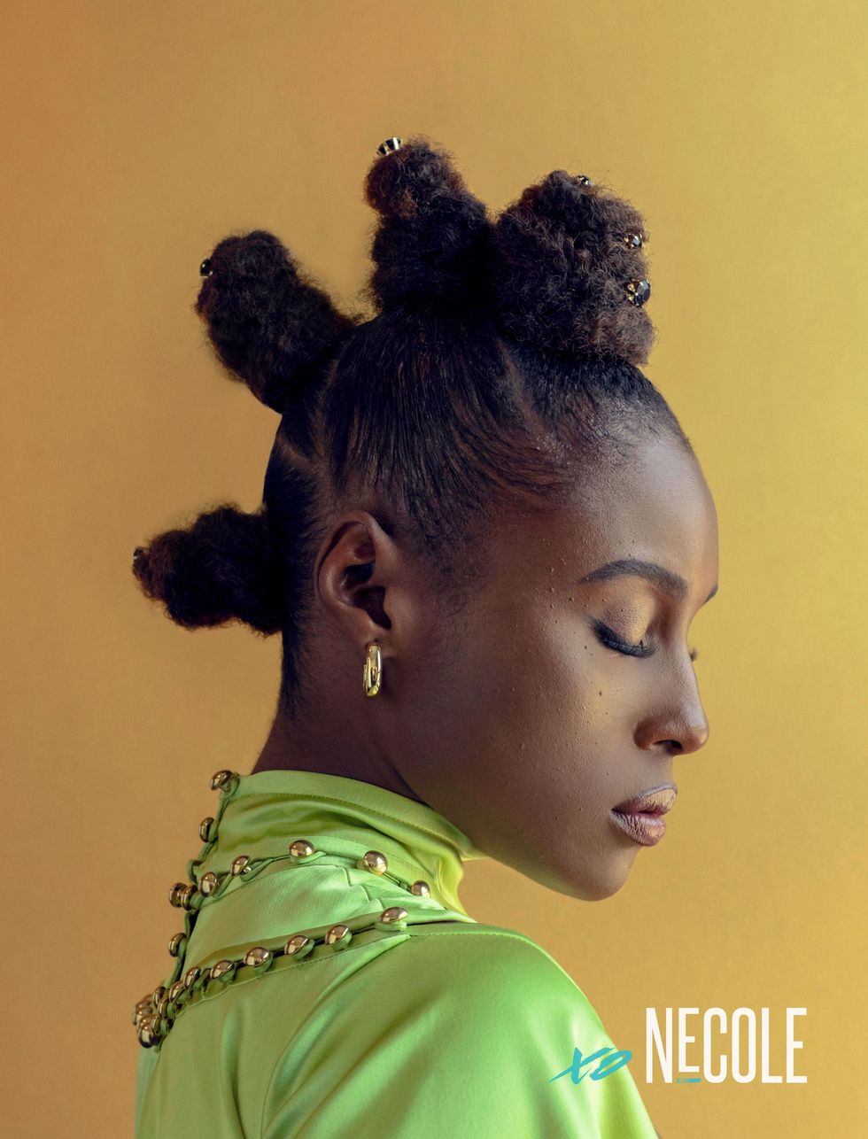 A Black woman, Issa Rae, has her hair in four Bantu knots in a mohawk style down her head. She looks down, eyes closed. Her lime green gown with gold embellishments covers her neck and shoulders. Her hoop earring are gold.