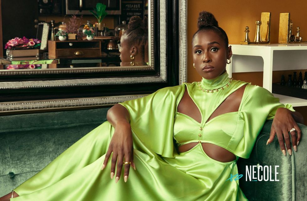A Black woman, Issa Rae, sits on a dark green velvet couch in a regal, lime green Prabal Gurung gown with cut outs on both sides of her chest and rib cage. The flowing short sleeves stop at her elbows. She rests her elbows on the back of the sofa and she stares directly into the camera. She's sitting in front of a mirror