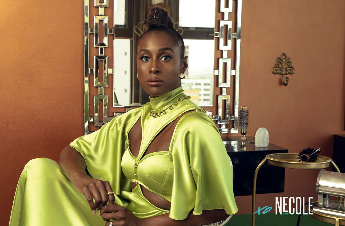 A Black woman, Issa Rae, sits in a salon chair in a regal, lime green Prabal Gurung gown with cut outs on both sides of her chest and rib cage. The flowing short sleeves stop at her elbows. She delicately holds her fingers in her lap and stares directly into the camera. She's sitting in front of a mirror, with hair brushes behind her on the counter and a hot press stove