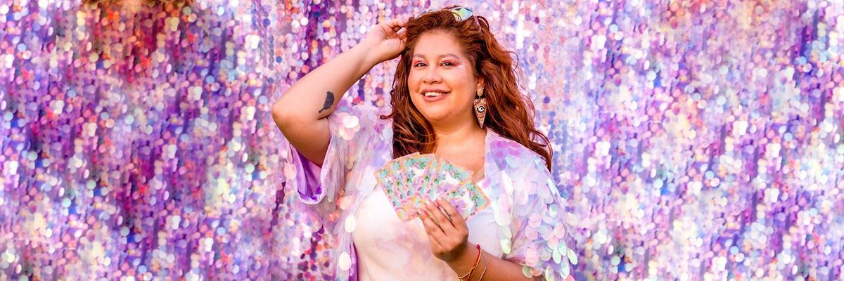 astrologer La Bruja del 305 stands with tarot cards fanned in her hand in front of purple toned large sequined wall