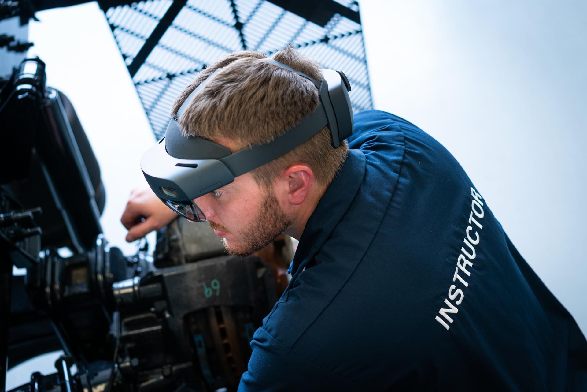 Penske instructor, wearing a blue uniform with the word instructor across the back in all caps, crouched down looking at a diesel truck engine. He is wearing mixed reality HoloLens Goggles for training purposes.